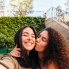 two young women pose for a selfie together as one kisses the other on the cheek, as they consider wh...